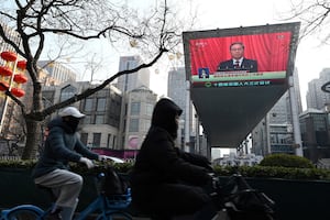 People ride past a screen showing live coverage of Chinese Premier Li Qiang speaking at the opening session of the National People’s Congress outside a shopping mall in Beijing on March 5, 2025.